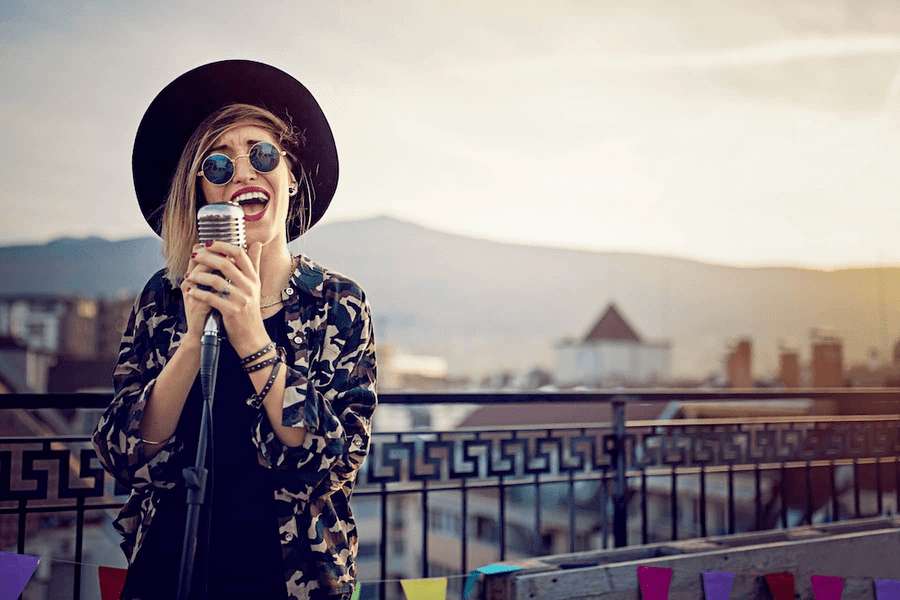 Une chanteuse et musicienne avec un chapeau et des lunettes de soleil se tient sur un toit, elle a un micro dans les mains.