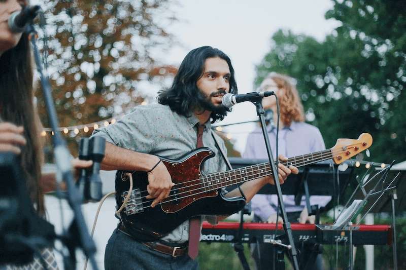 Un groupe de musique se tient debout sur scène, le guitariste et chanteur se tient devant le micro, accompagné d'une autre chanteuse et d'un pianiste lors d'un concert en plein air.