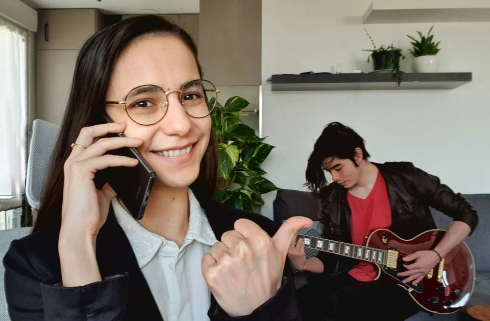 Une jeune bookeuse de Linkaband est au téléphone dans un salon pendant que, derrière elle, l’un de ses artistes, un jeune guitariste, joue de la guitare, assis sur le canapé.