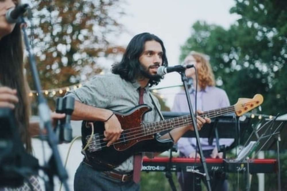 Un groupe de musique se tient debout sur scène, le guitariste et chanteur se tient devant le micro, accompagné d'une autre chanteuse et d'un pianiste lors d'un concert en plein air.